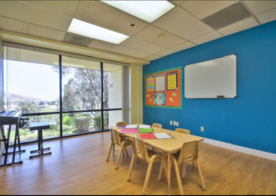A large classroom with a group table, a electronic piano, and a large window.