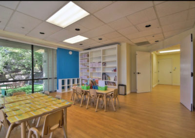 A large room with group tables, a large bookshelf, and classroom supplies.